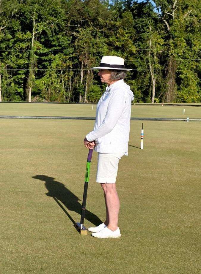 Person in white attire holding a mallet on a croquet lawn, wearing a hat, with trees in the background.