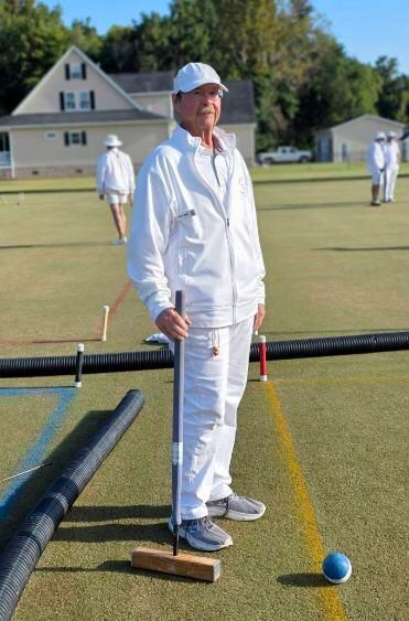 Man in white croquet attire on green field, holding mallet, ball nearby.