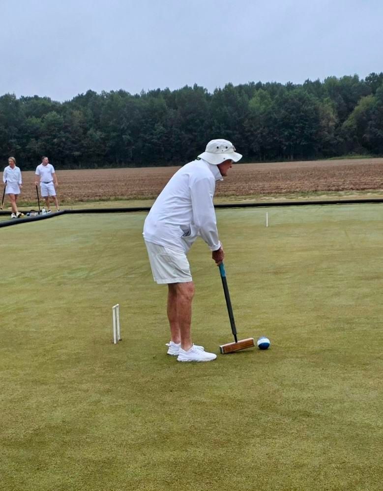 Man in white plays croquet on a green lawn, aiming mallet at ball. Two others watch in background.