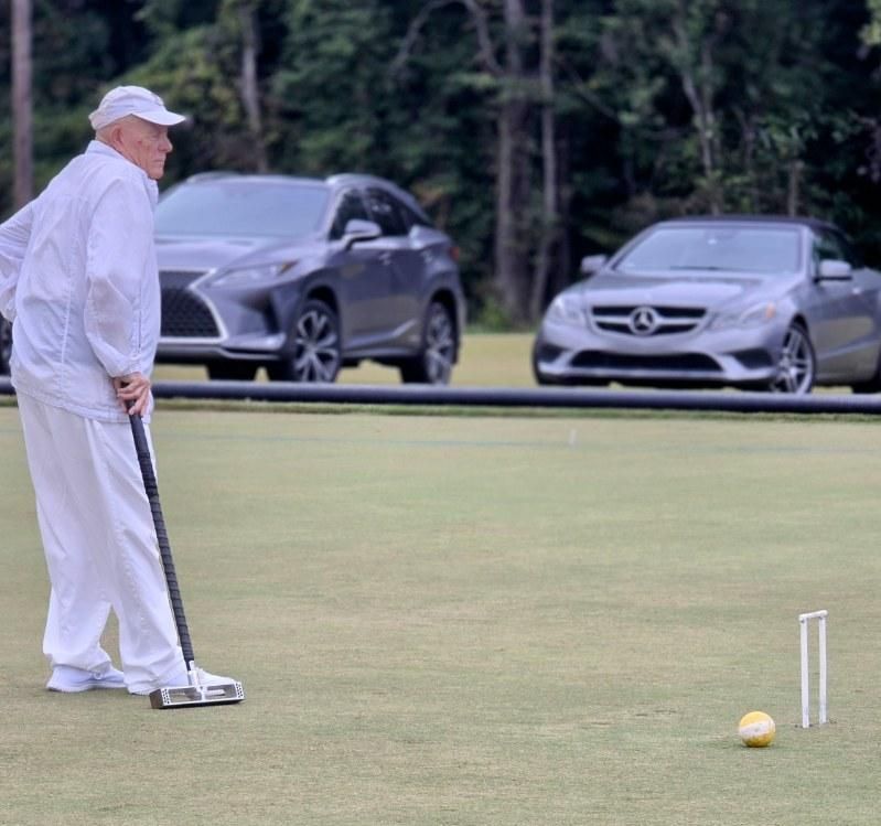 Man in white playing croquet on a green lawn, two cars parked in background.
