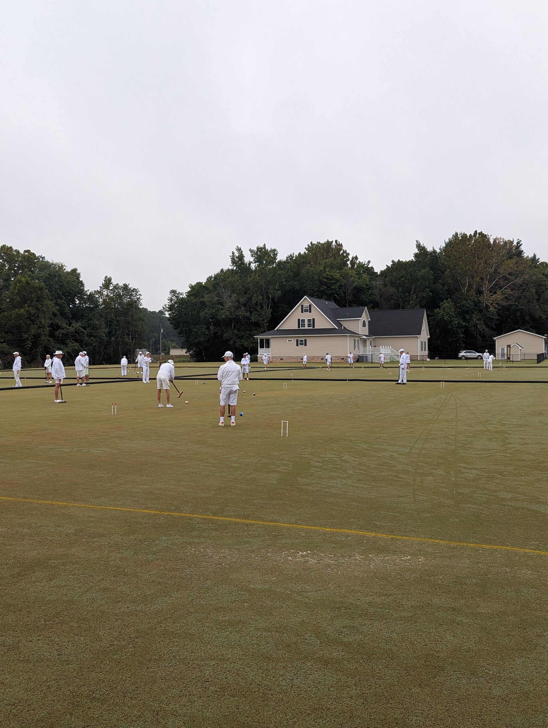 People in white play croquet on a green field, a building in the background. Overcast sky.