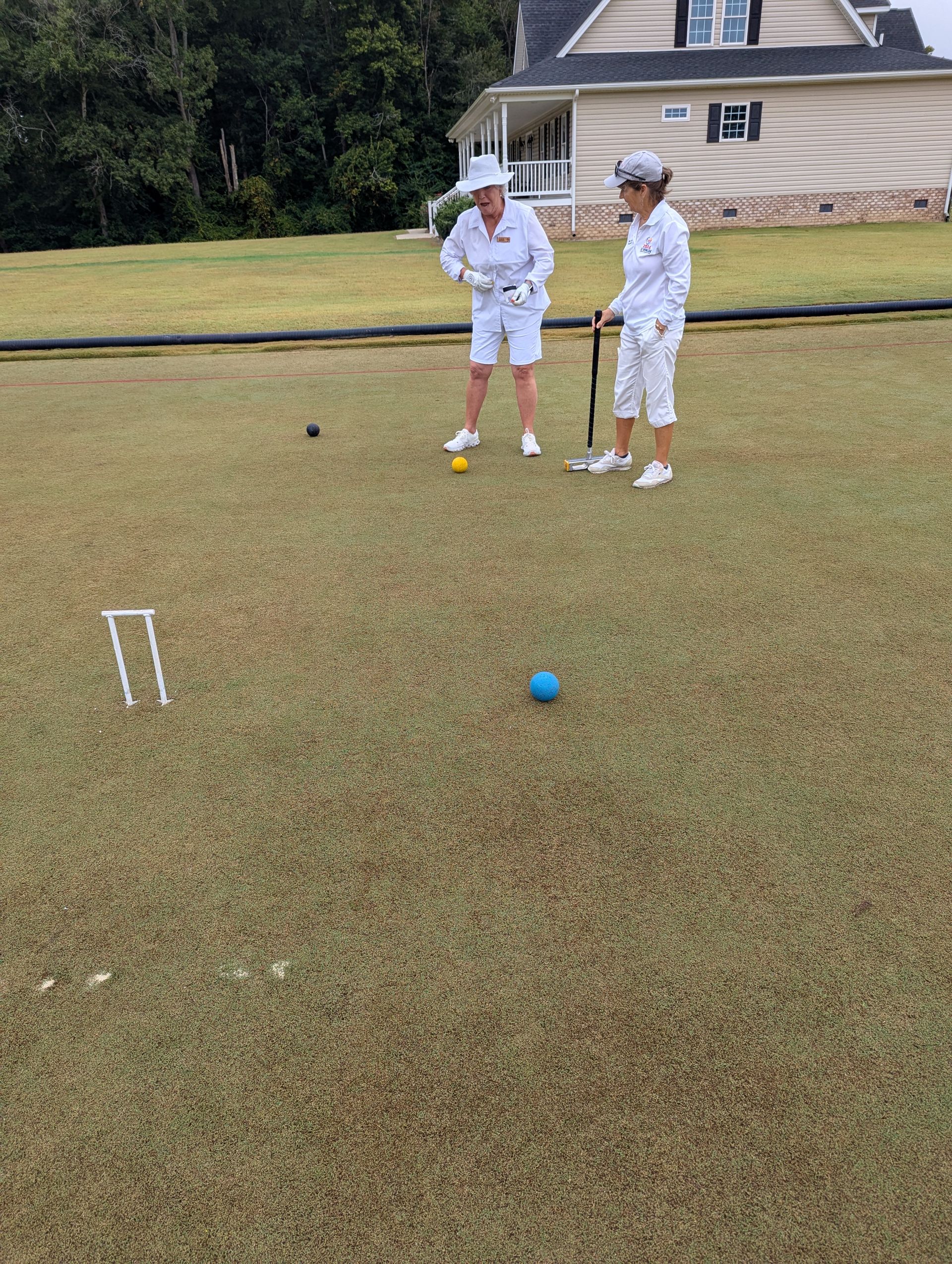 Two people in white playing croquet on a green lawn, house in the background.
