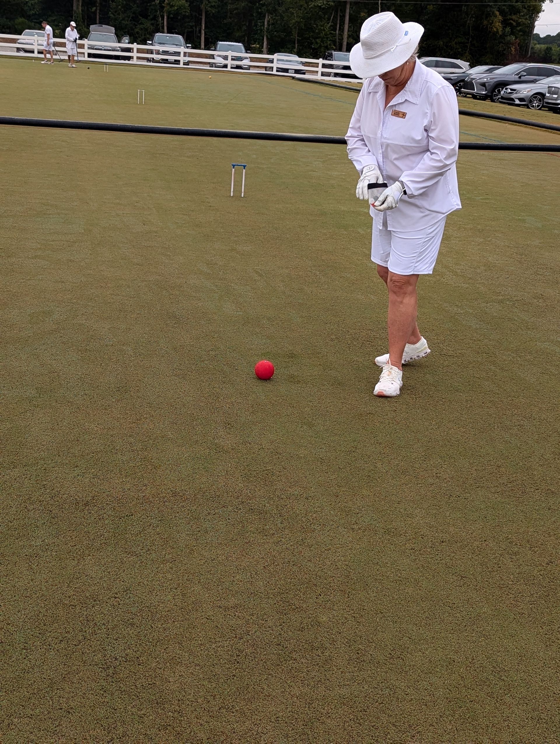 A person in white playing croquet on a green lawn, aiming at a red ball with a mallet.