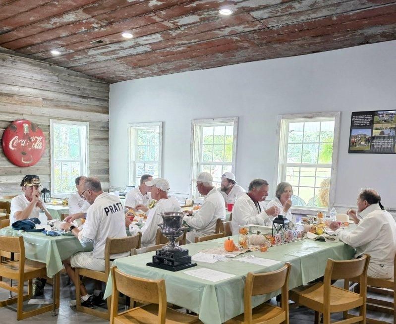 People in white attire at a luncheon inside a rustic room with long tables, enjoying a meal.