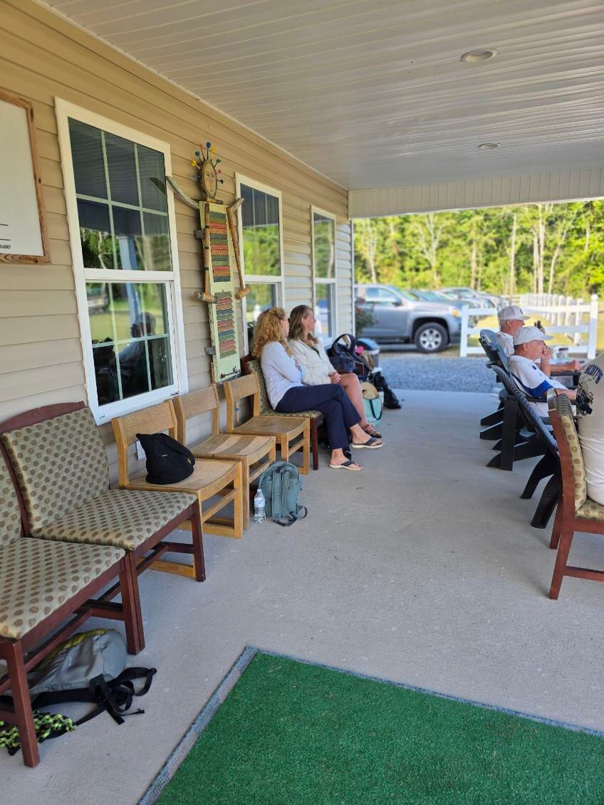 People sitting on benches and chairs on a covered porch, near a grassy area and parking lot.
