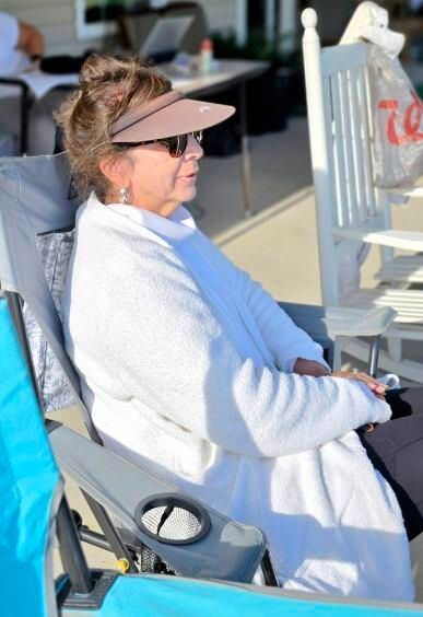 Woman in sunglasses and visor, wearing a white robe, seated in a chair outdoors.