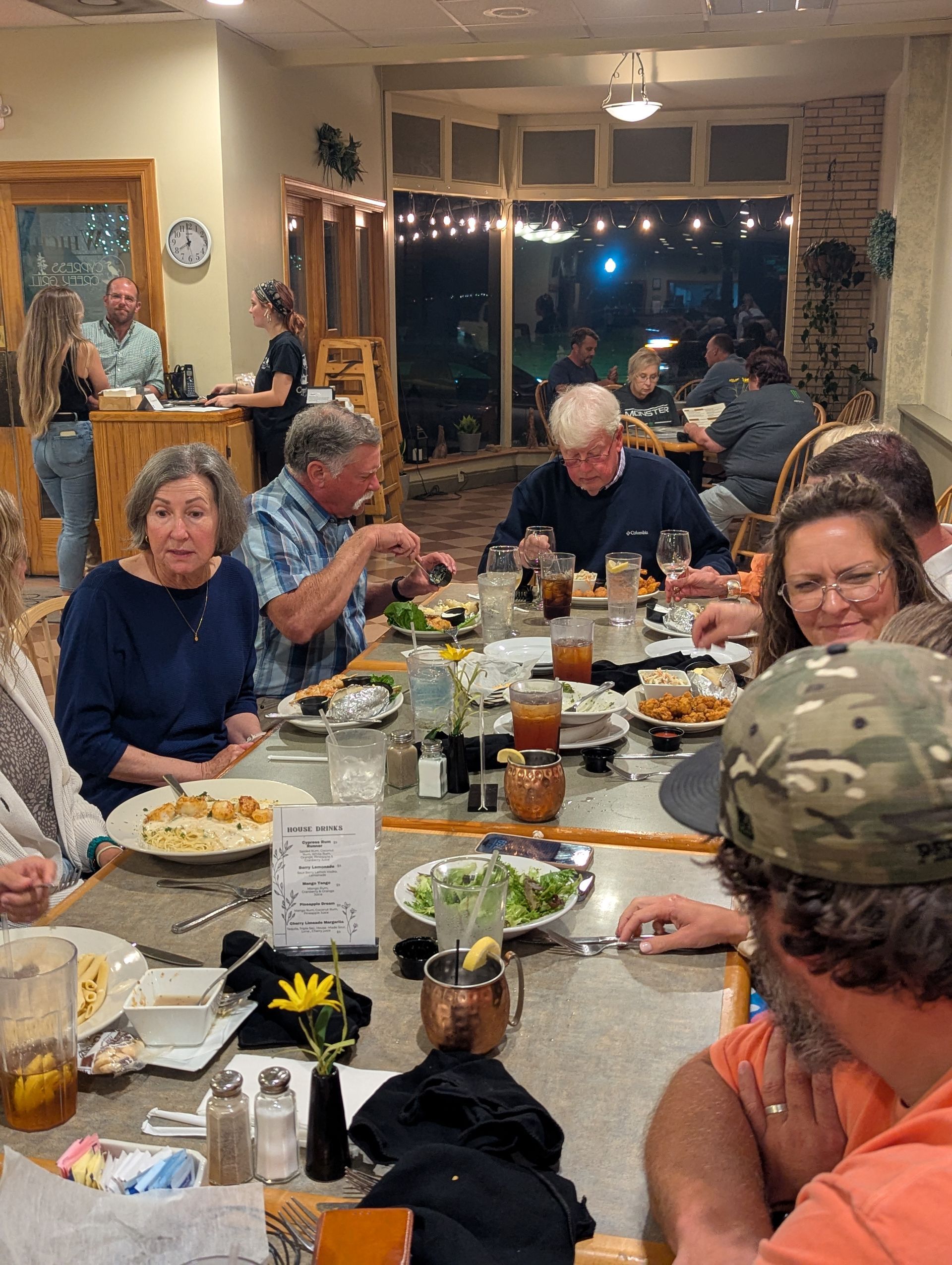 People seated at tables in a restaurant, eating and talking. Other people are in the background, by a bar.