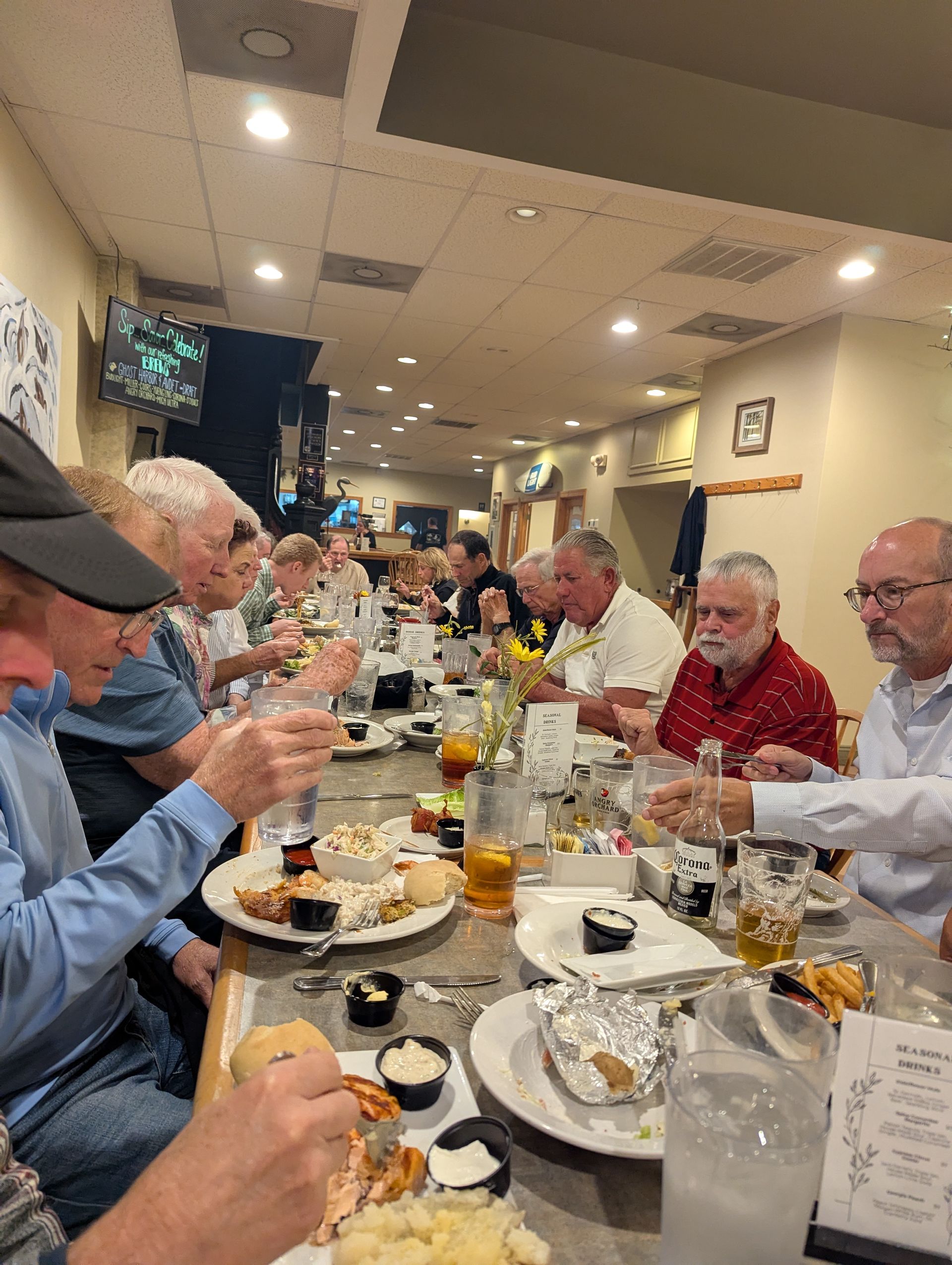 People dining at a restaurant, seated at a long table. Plates of food and drinks are visible.