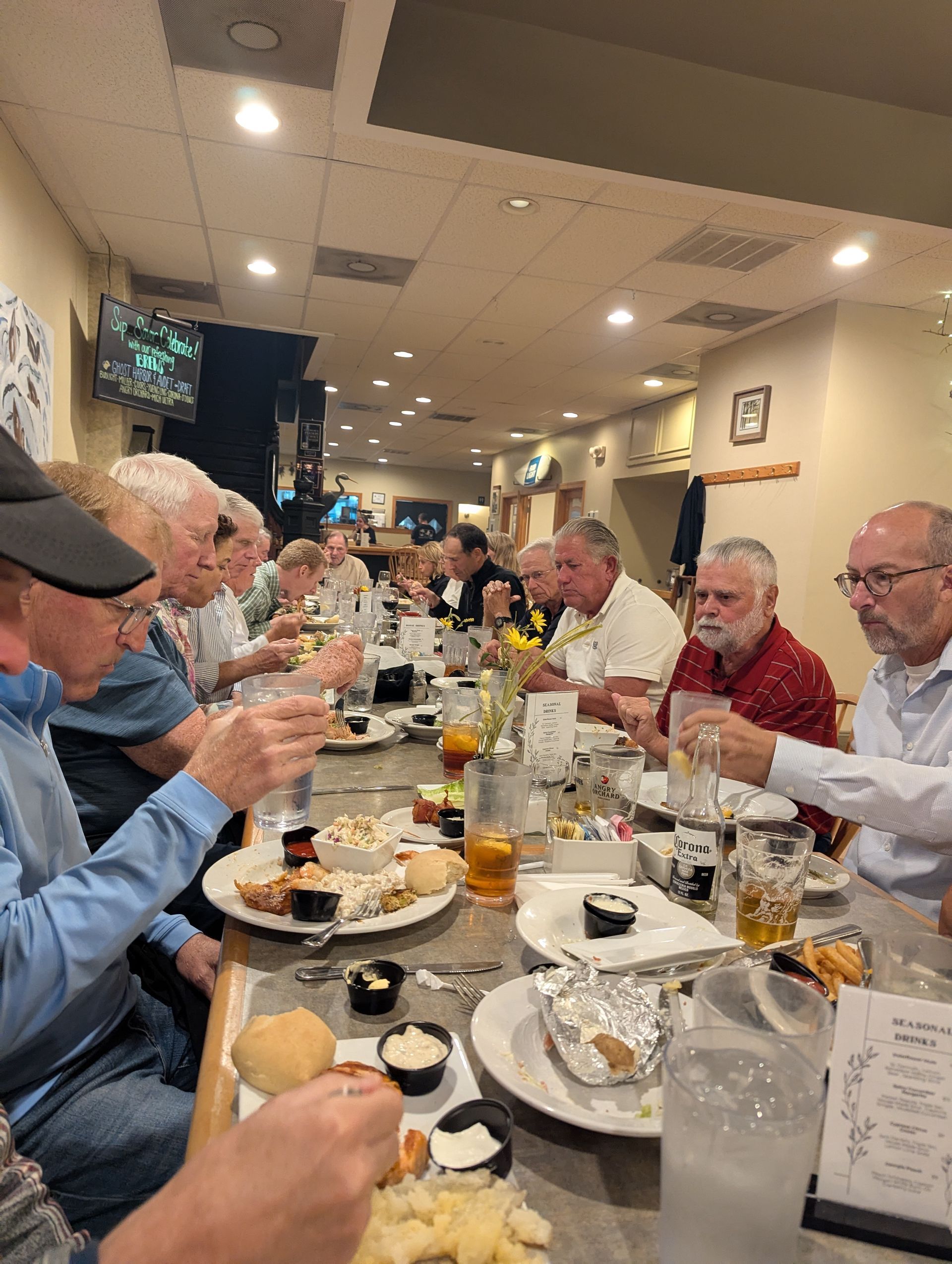 People seated at long tables eating a meal at a restaurant. Plates of food and drinks are on the table.