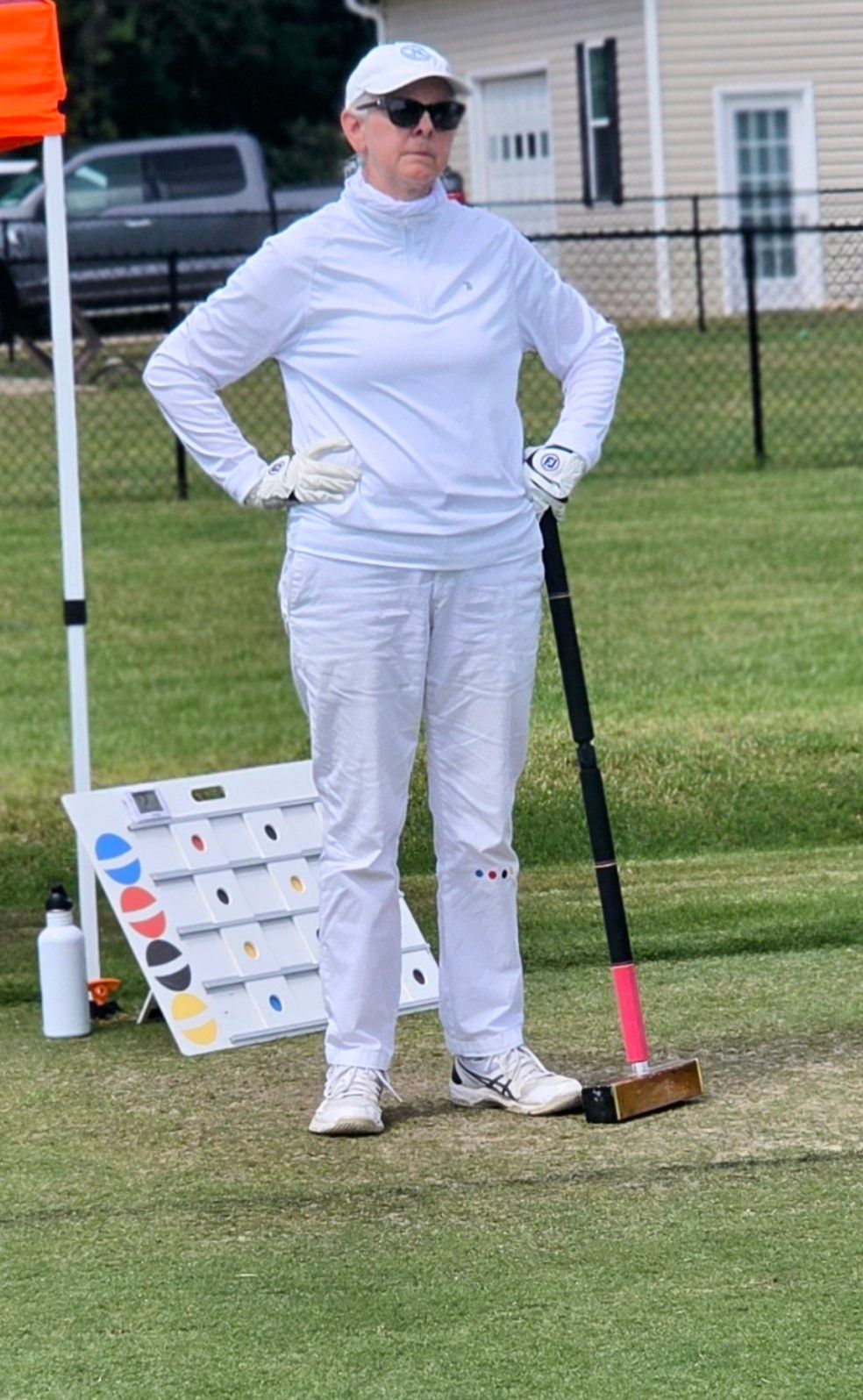 Person in white outfit, sunglasses, and hat stands with mallet on a croquet lawn.