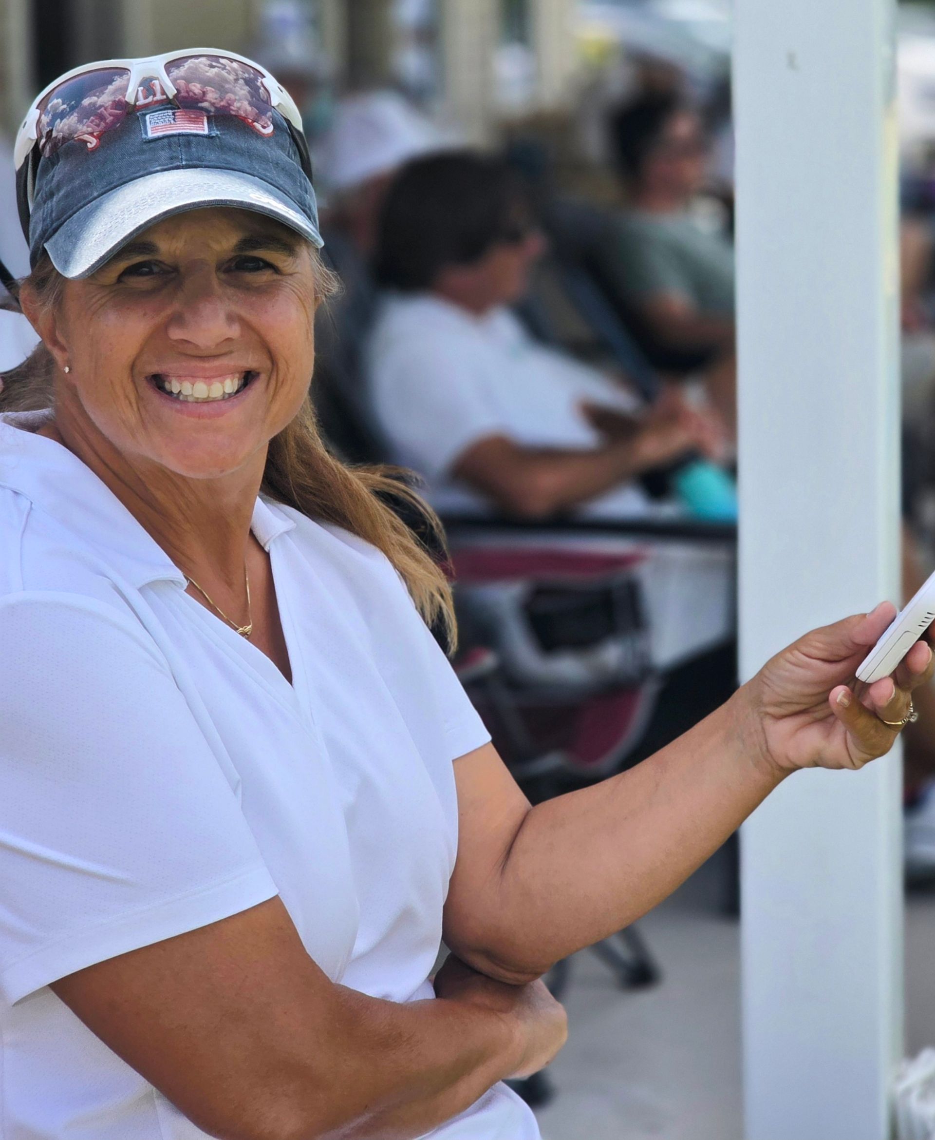 Woman in white shirt and denim cap smiles while holding a phone outdoors.