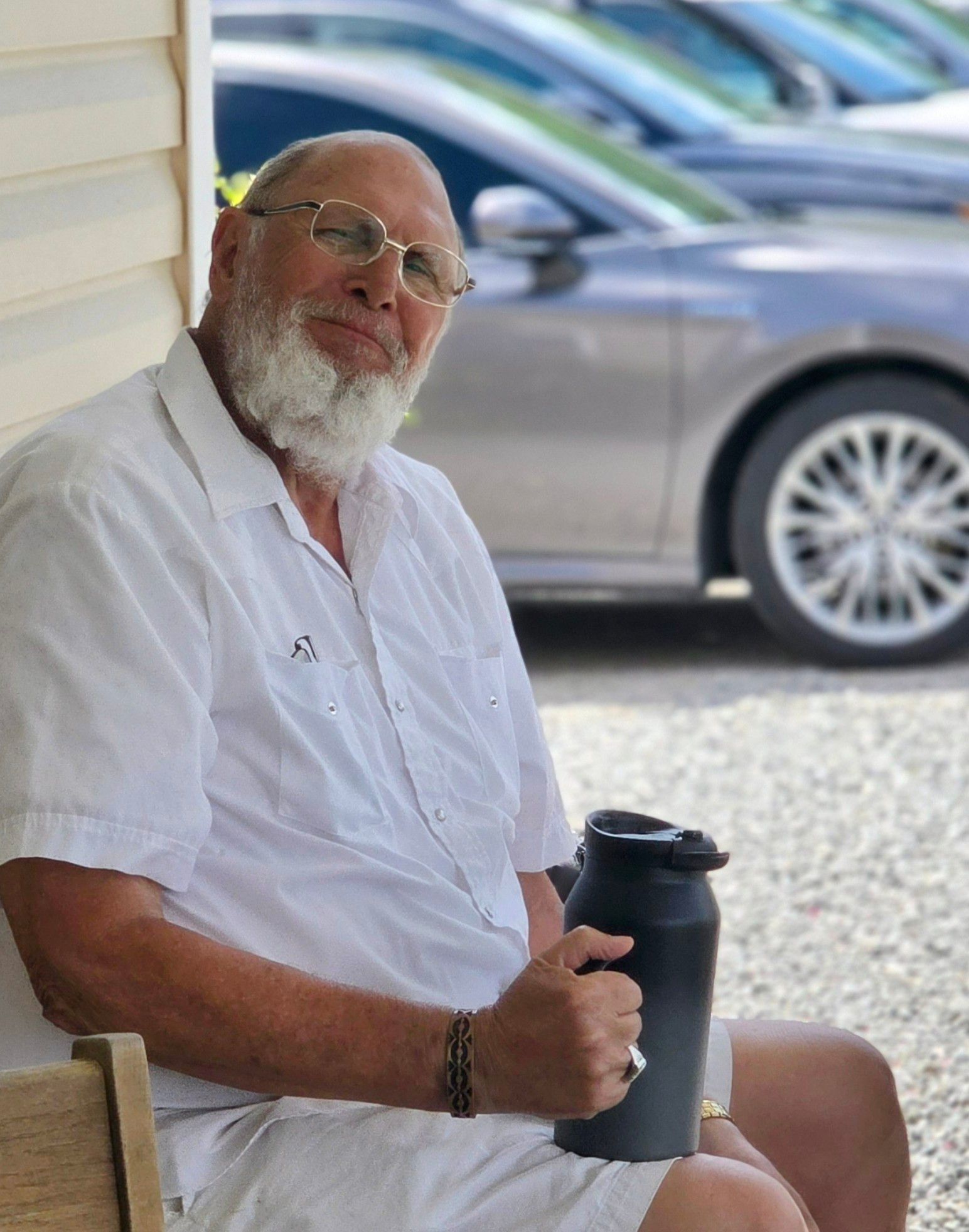 Man with a white beard, glasses, and white shirt sits holding a black tumbler. Cars in background.