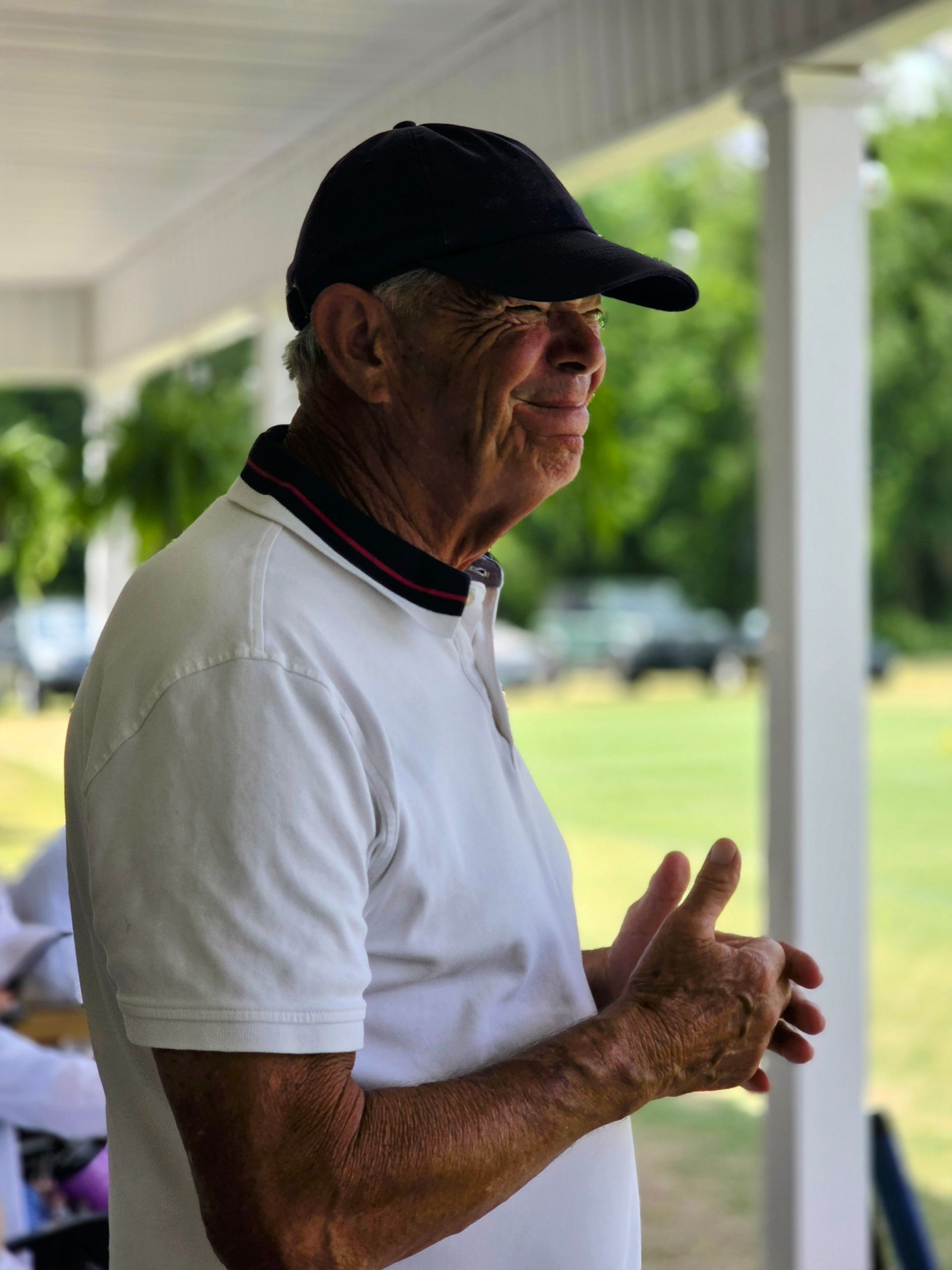 Man in a white polo shirt and black cap gestures with hands, standing outdoors near a support column.