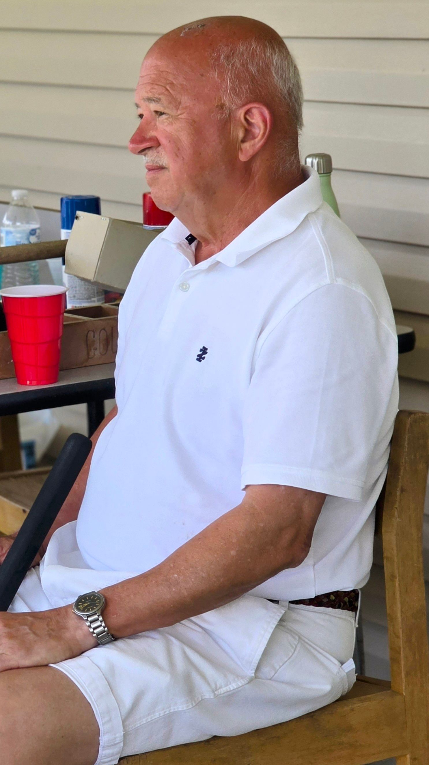 Man in white polo shirt and shorts sits on a wooden bench, outside.