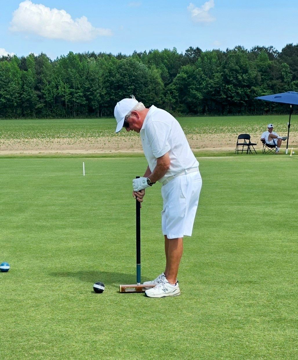 Man in white playing croquet on a green lawn, preparing to strike the ball with his mallet.