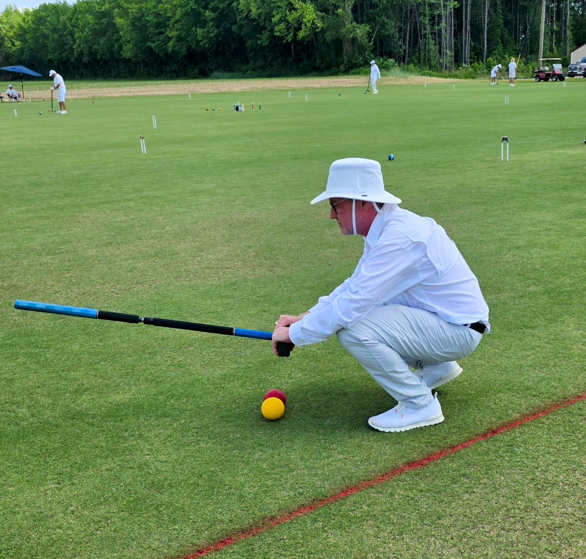 Man in white, playing croquet on a green lawn, aiming mallet at ball. Other players in background.