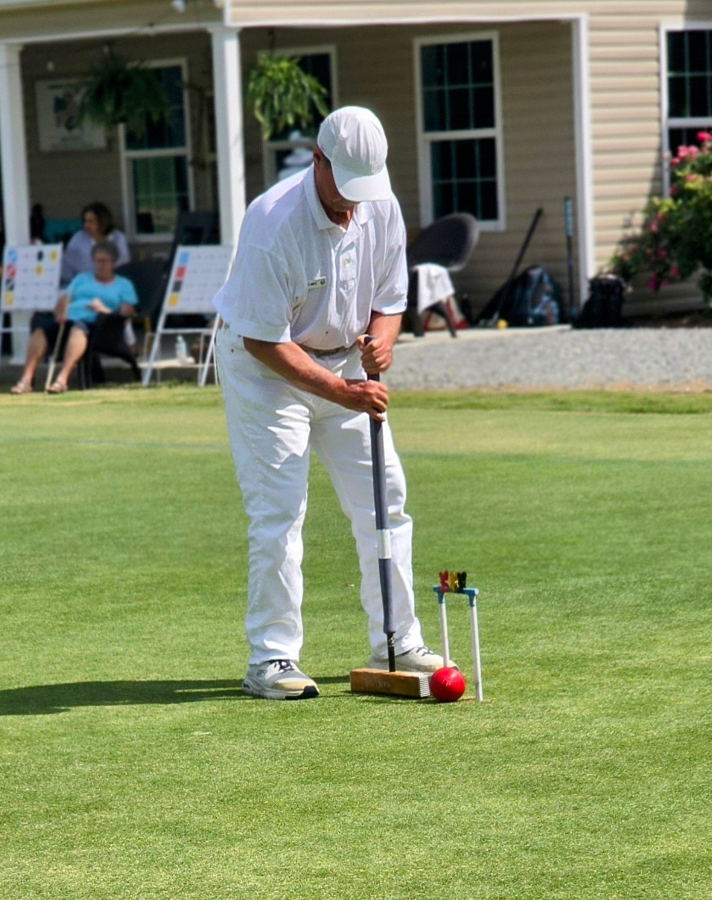 Man in white attire playing croquet on a green lawn, aiming mallet at the red ball.