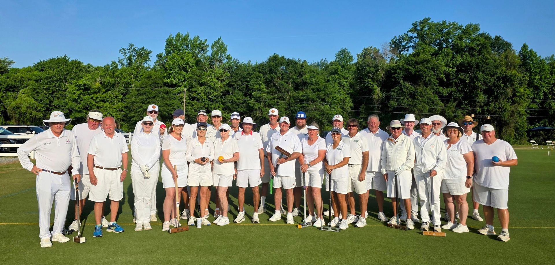 A group of people wearing white outfits and hats on a green field. Trees are in the background.