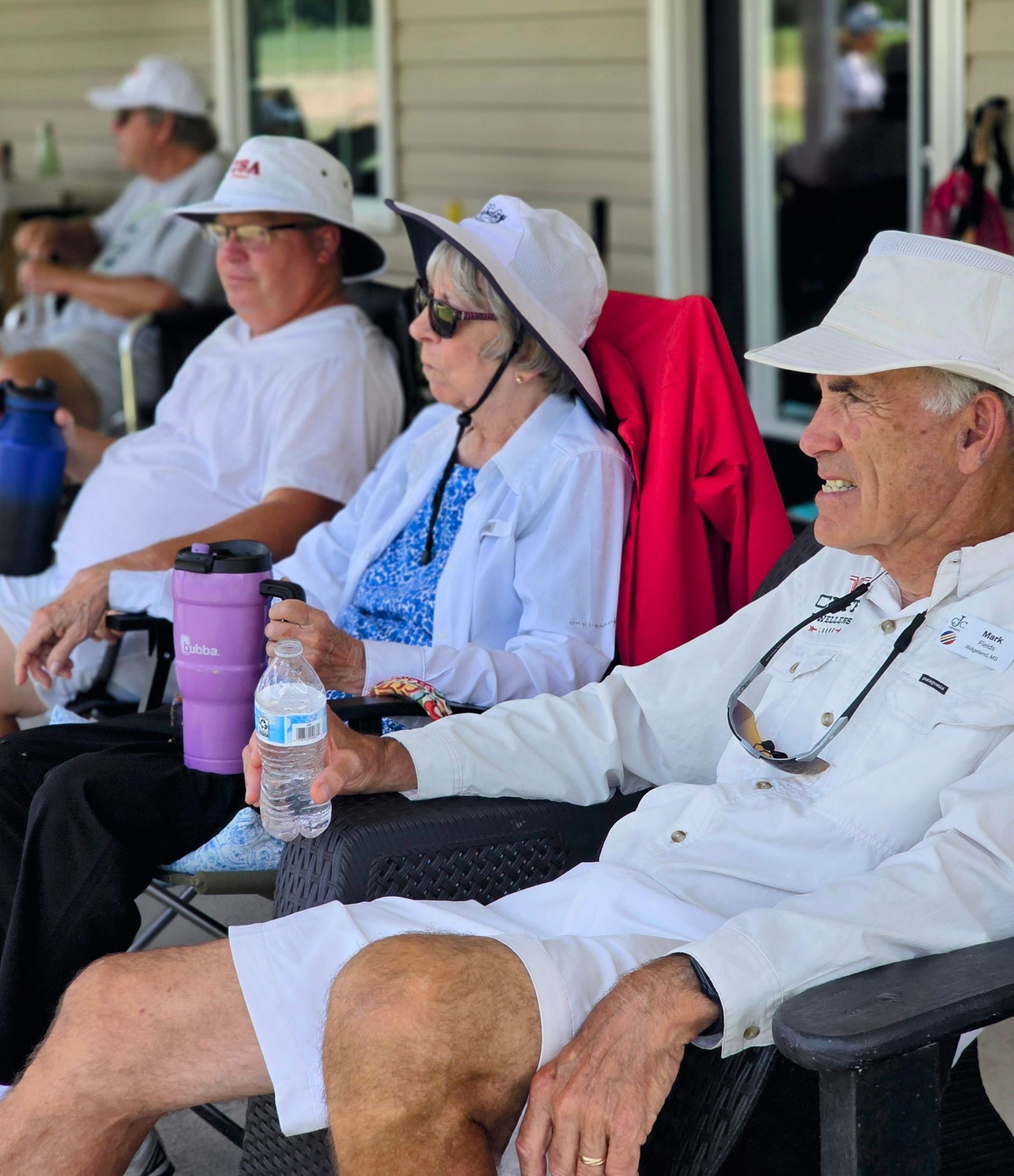 People seated outside, wearing white hats and shirts. One person holds a water bottle.
