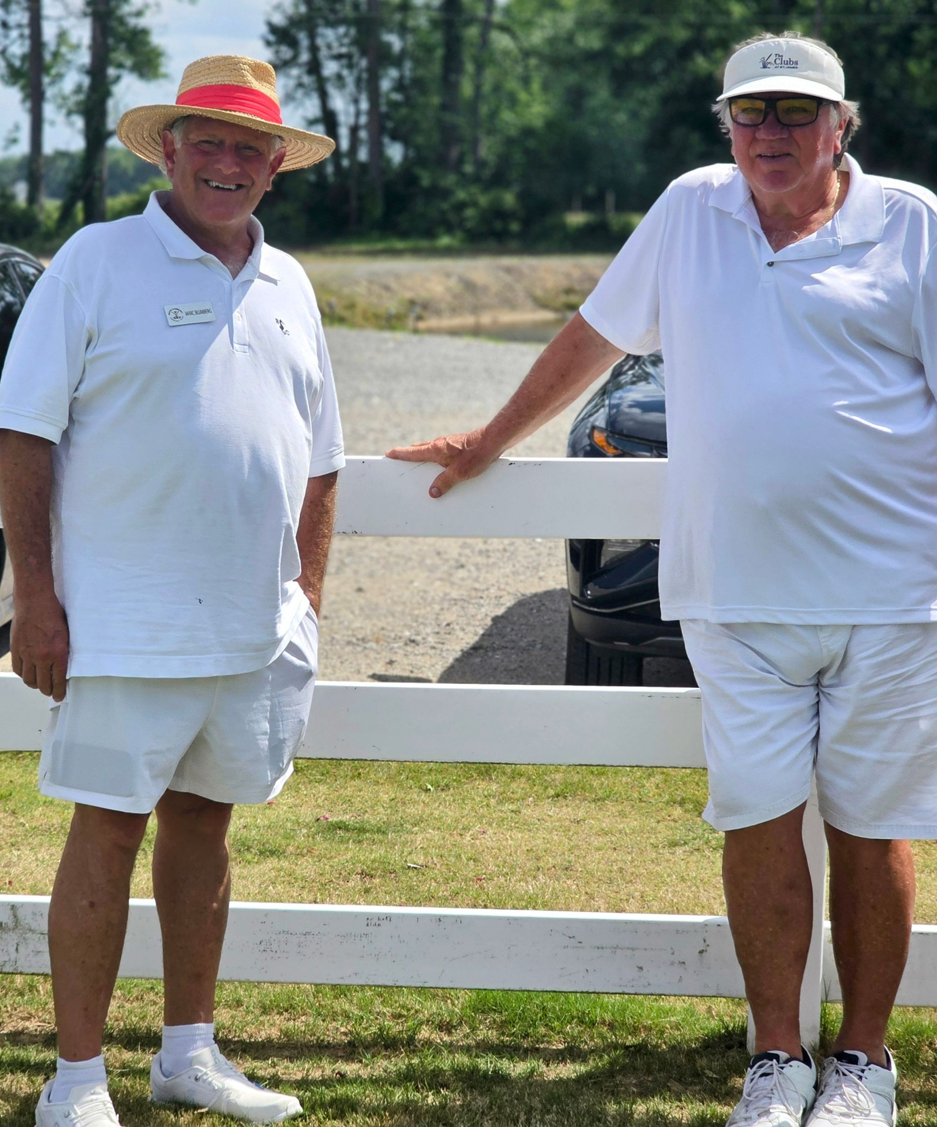 Two men in white outfits stand by a white fence outdoors on a sunny day. One wears a straw hat.