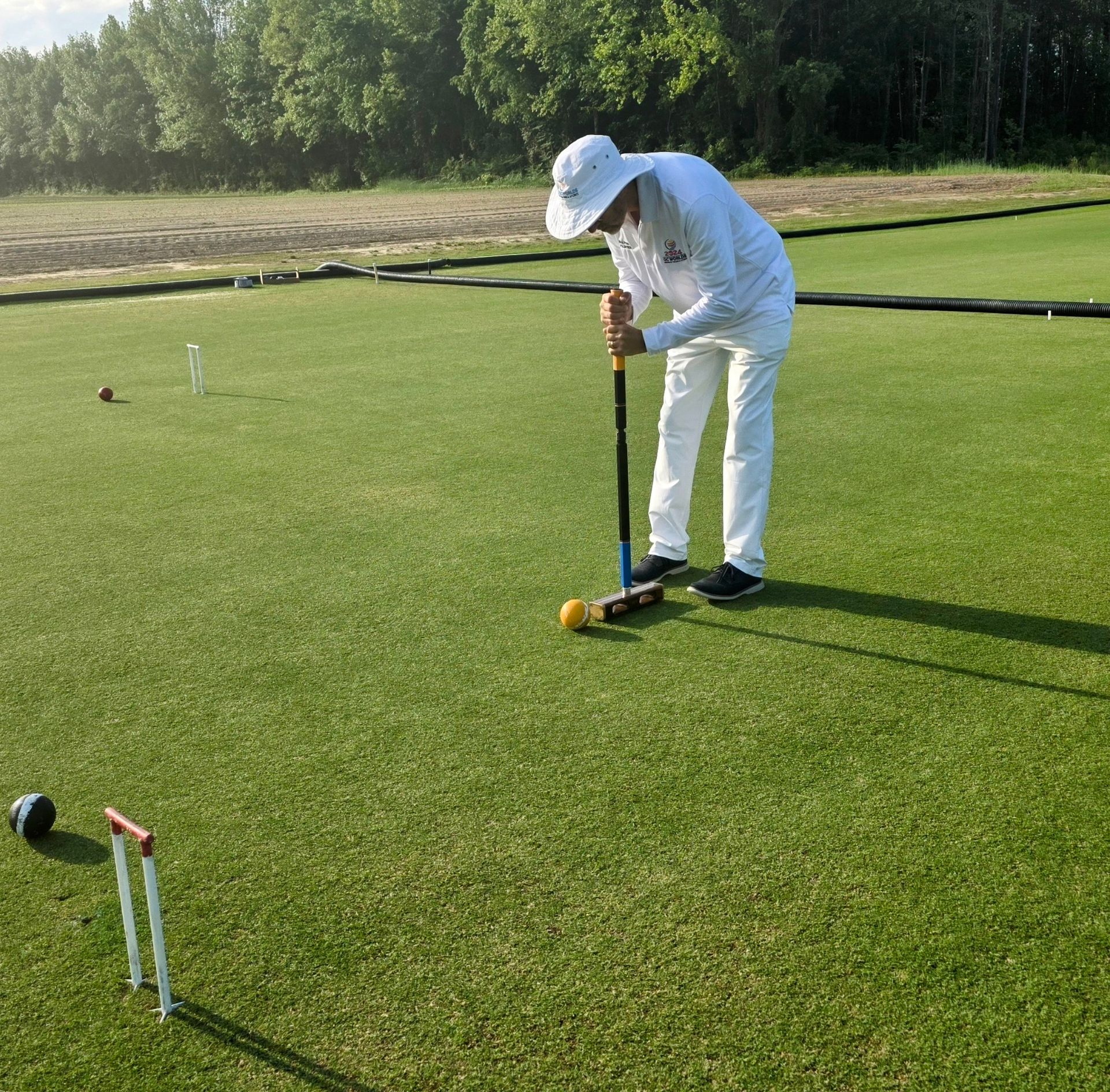 Person in white playing croquet on a green lawn, aiming mallet at a yellow ball near a hoop.