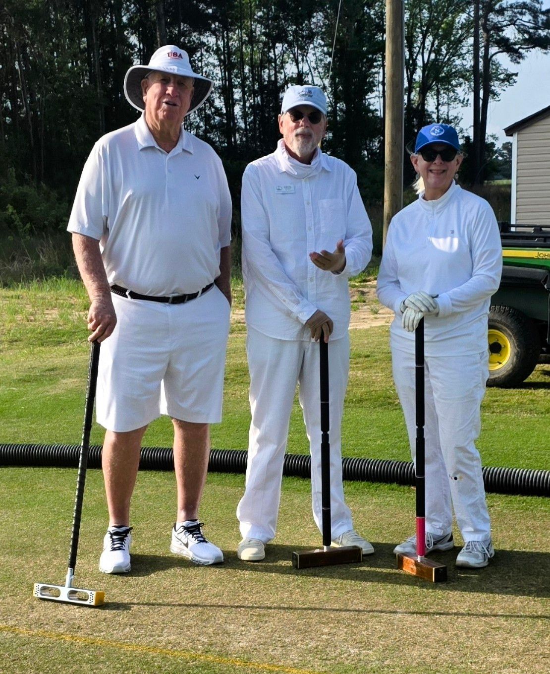 Three people in white clothing standing on a croquet lawn, holding mallets.