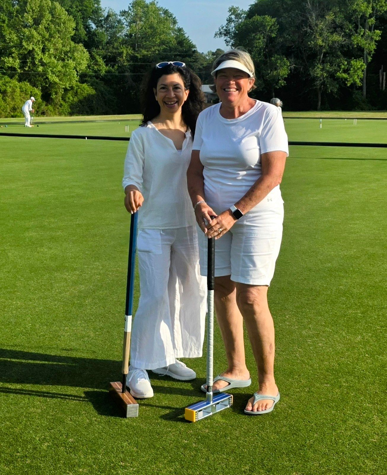 Two women in white on a croquet lawn, holding mallets. One is smiling. Green grass and trees in background.