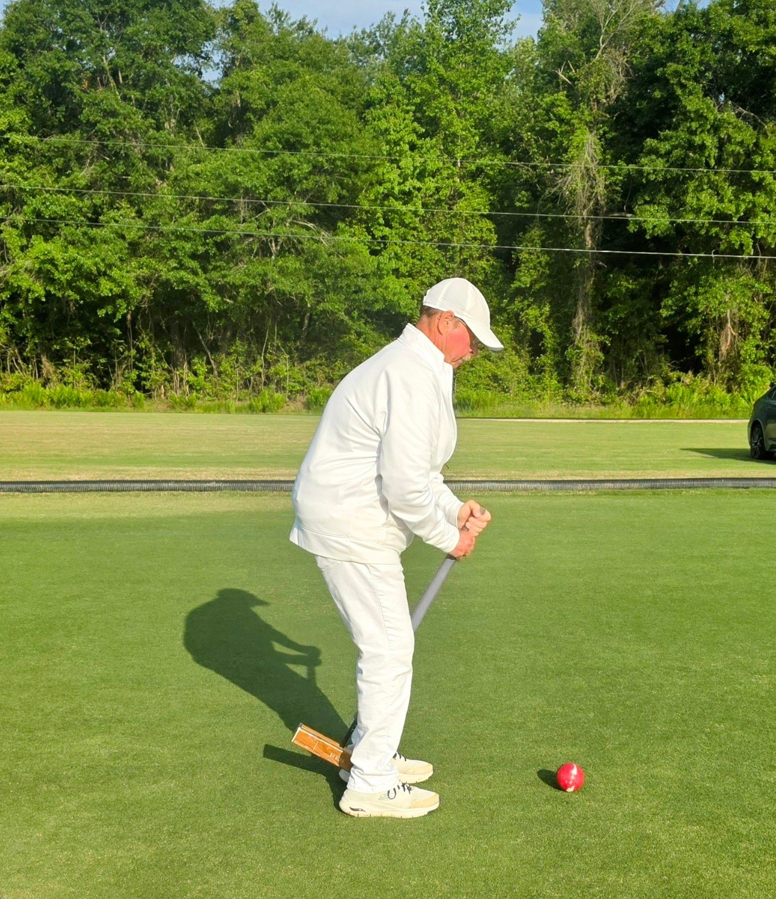 Person in white playing croquet on a green lawn; red ball and mallet present.