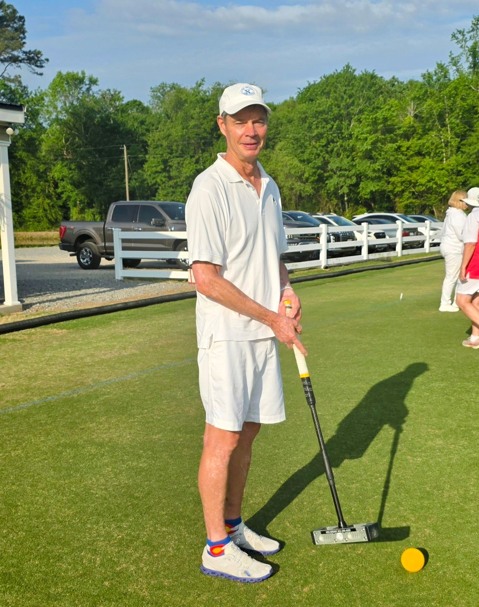 Man in white outfit stands on a croquet lawn, holding a mallet. Yellow ball nearby.