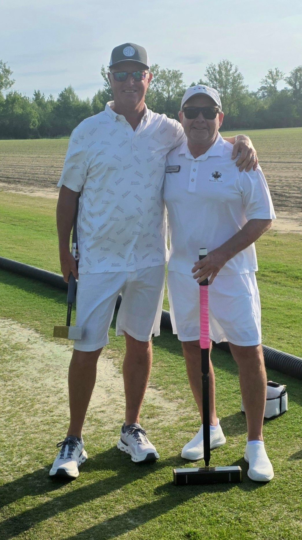 Two men in white outfits pose with croquet mallets on a green field.