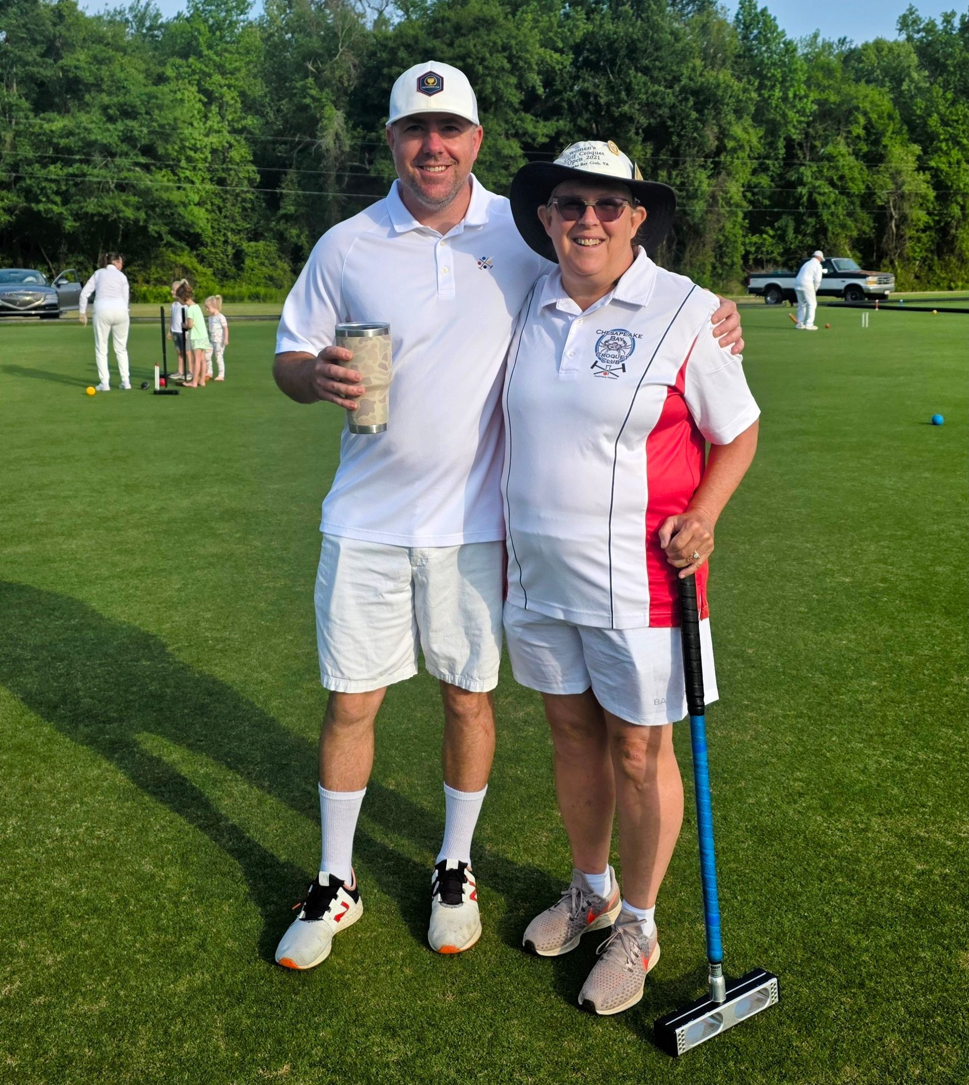 Two people on croquet lawn; one holding trophy, other a mallet. Both in white attire. Green field, sunny.