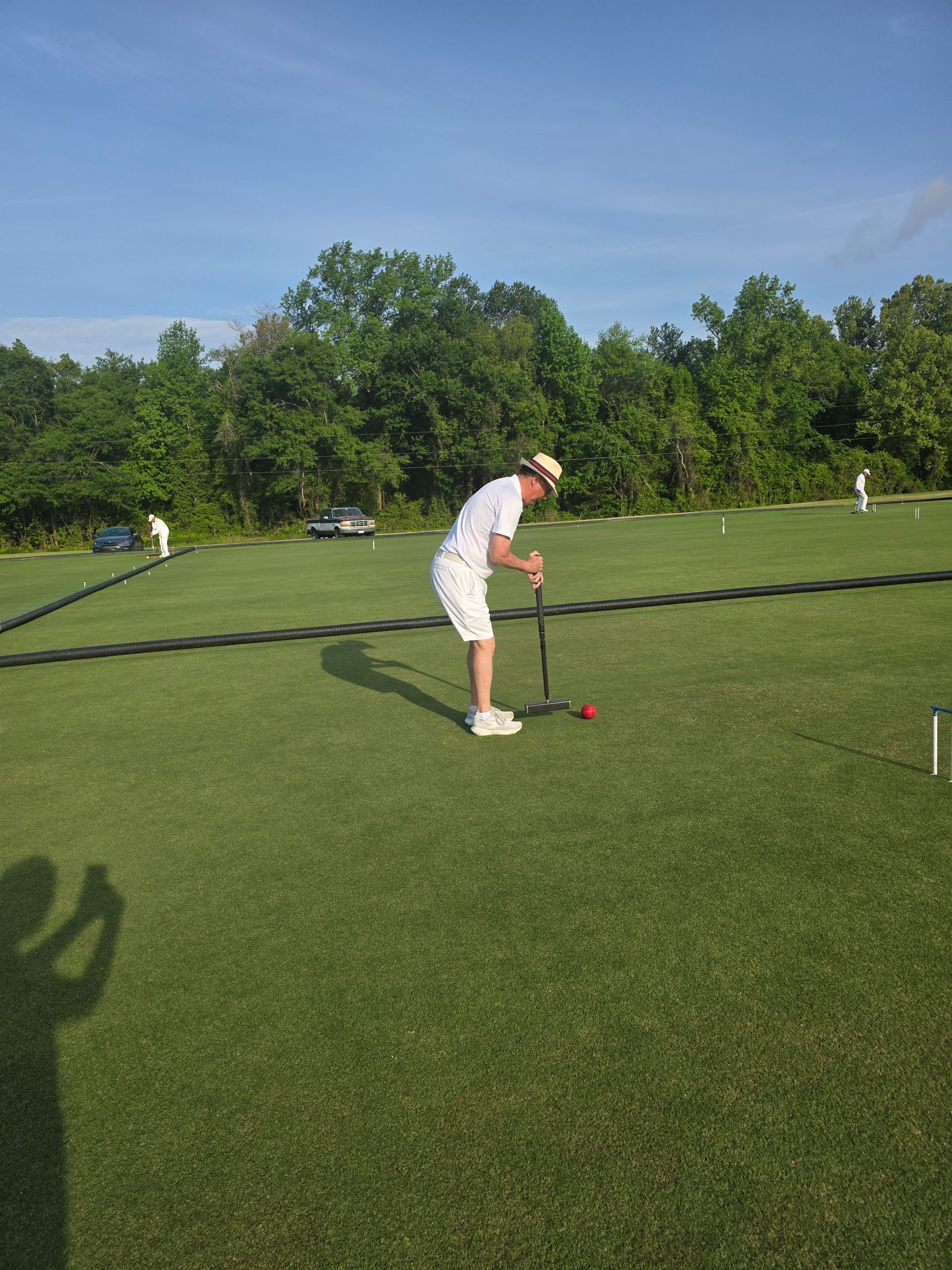 Man in white plays croquet on a green lawn with trees in the background under a blue sky.