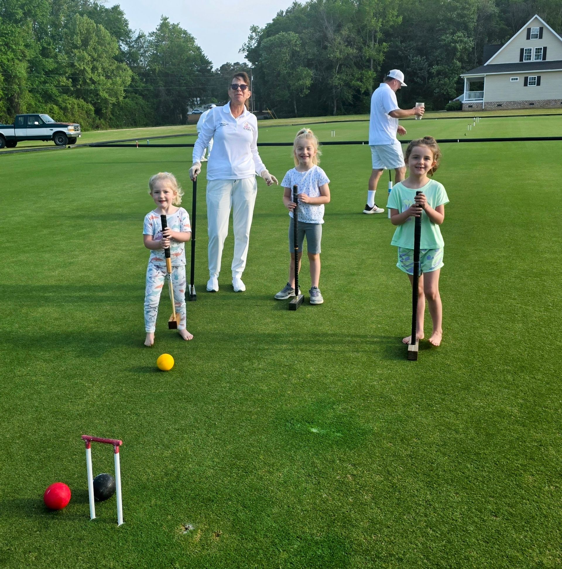 Family playing croquet on a green lawn. Adults and children hold mallets; a yellow ball is in play.