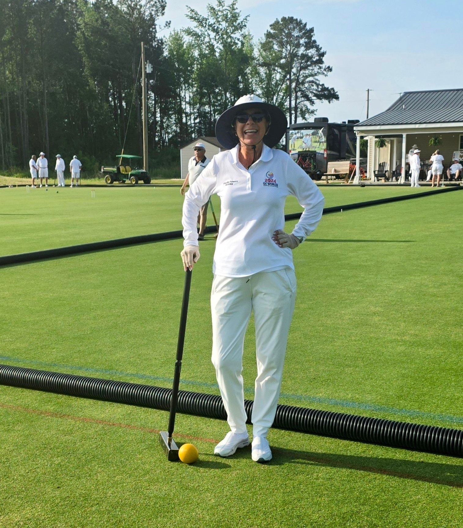 Woman in white outfit with mallet on a green lawn, other players in the background.