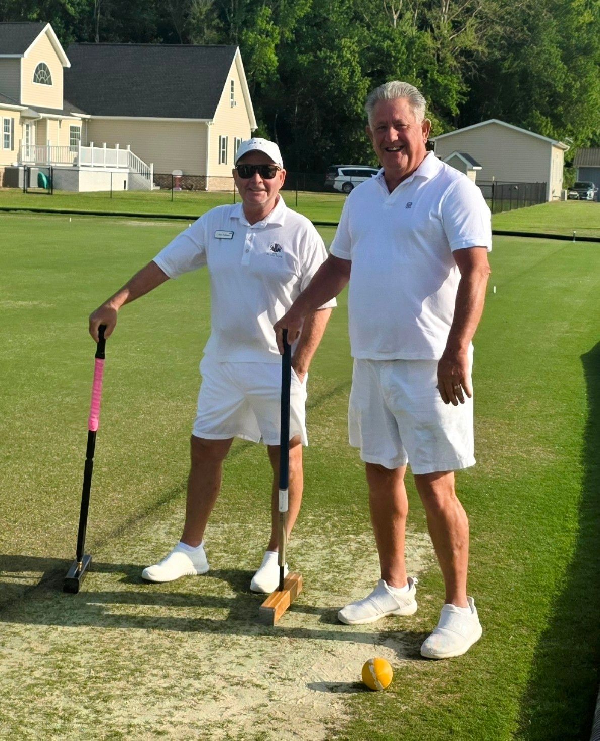 Two men in white outfits stand on a green lawn, holding croquet mallets.