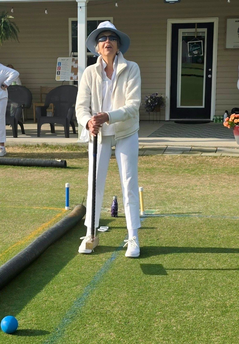 Woman in white playing croquet on a green lawn, holding mallet, sunny outdoor setting.