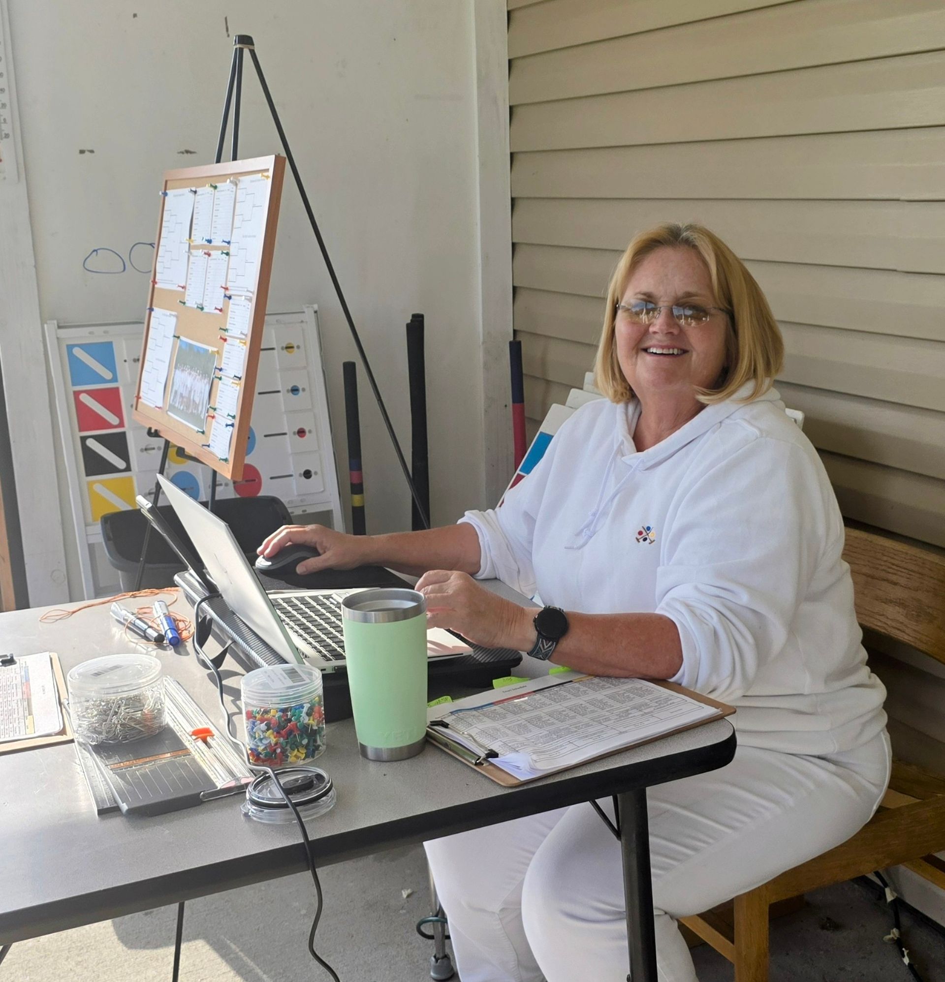 Woman smiling, working on laptop at outdoor table with paperwork, drink, and whiteboard.