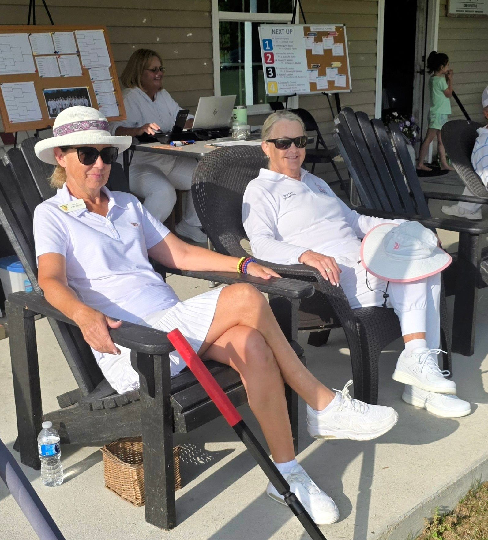 Two women in white outfits relax in chairs on a patio; a third person works at a laptop nearby.