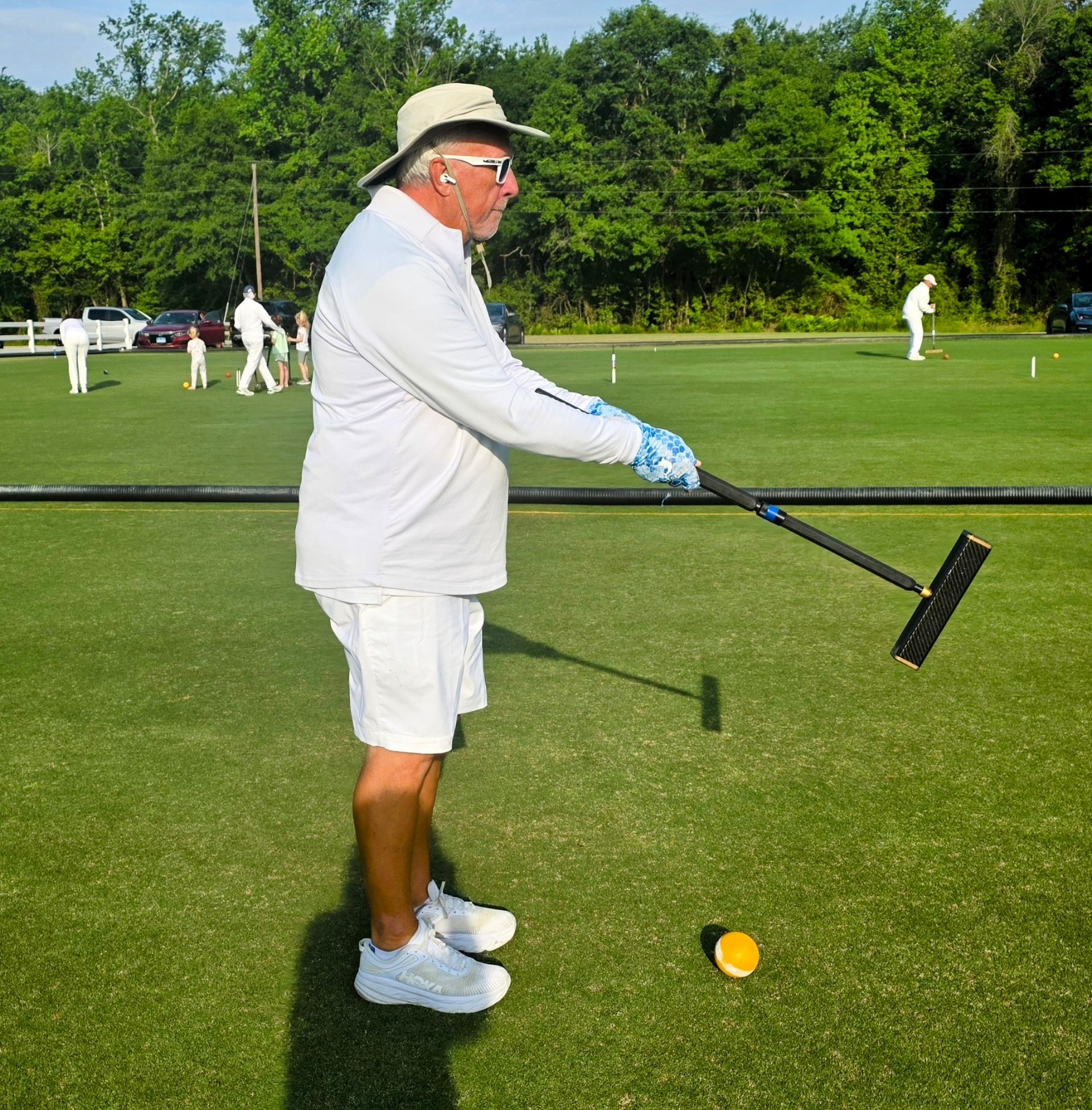 Man in white playing croquet on a green lawn; holding mallet, yellow ball nearby.