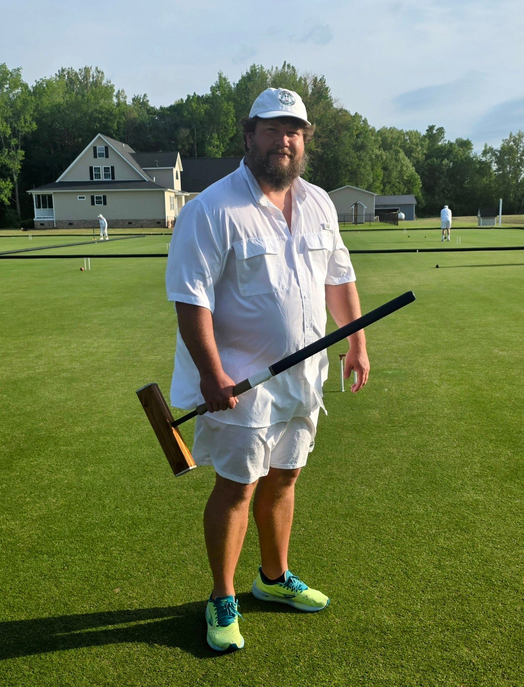 Man in white holding a croquet mallet on a green lawn, near a building, wearing a white hat and shorts.