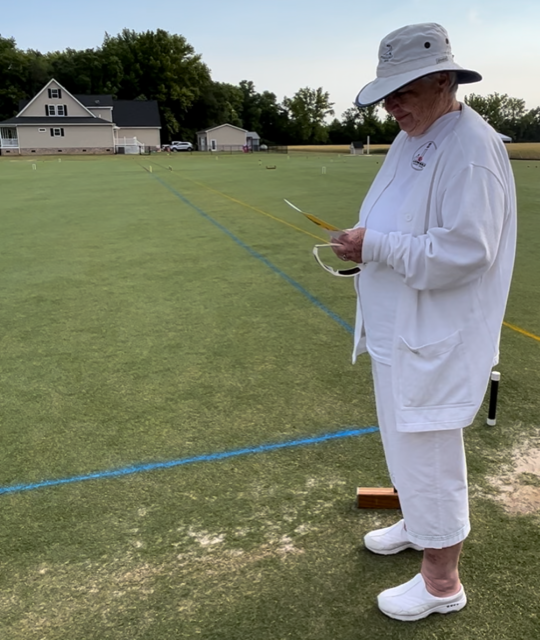 Person in white clothing and hat on a croquet lawn, looking at a scorecard.