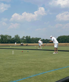 People playing croquet on a green field under a blue sky.