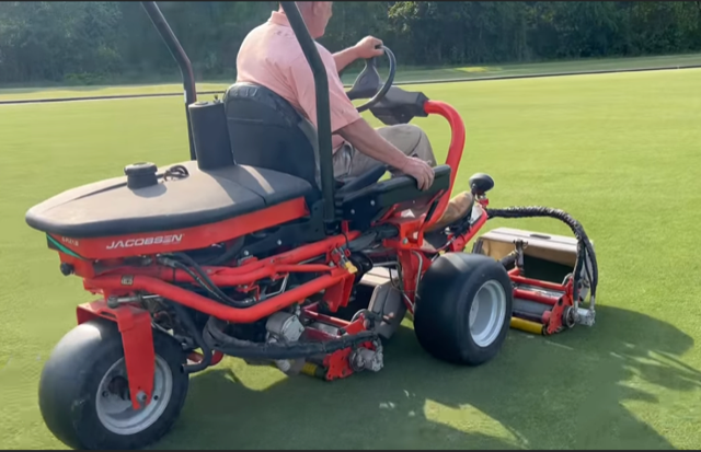 Person driving a red Jacobsen greens mower on a green lawn.