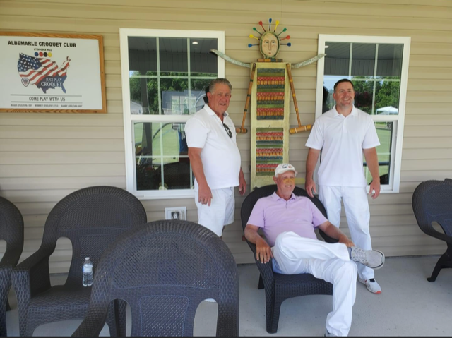 Three men in white clothing on a porch. One seated in a chair, two standing. Building in the background.