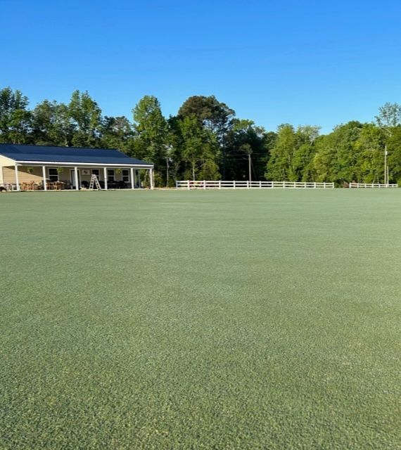 A green lawn with a white fence, building, and trees against a blue sky.