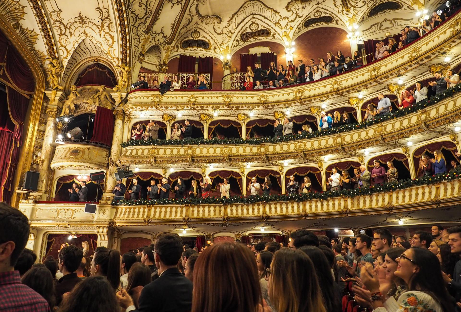 Ornate theater interior filled with people watching a performance. Golden balconies and ceiling.