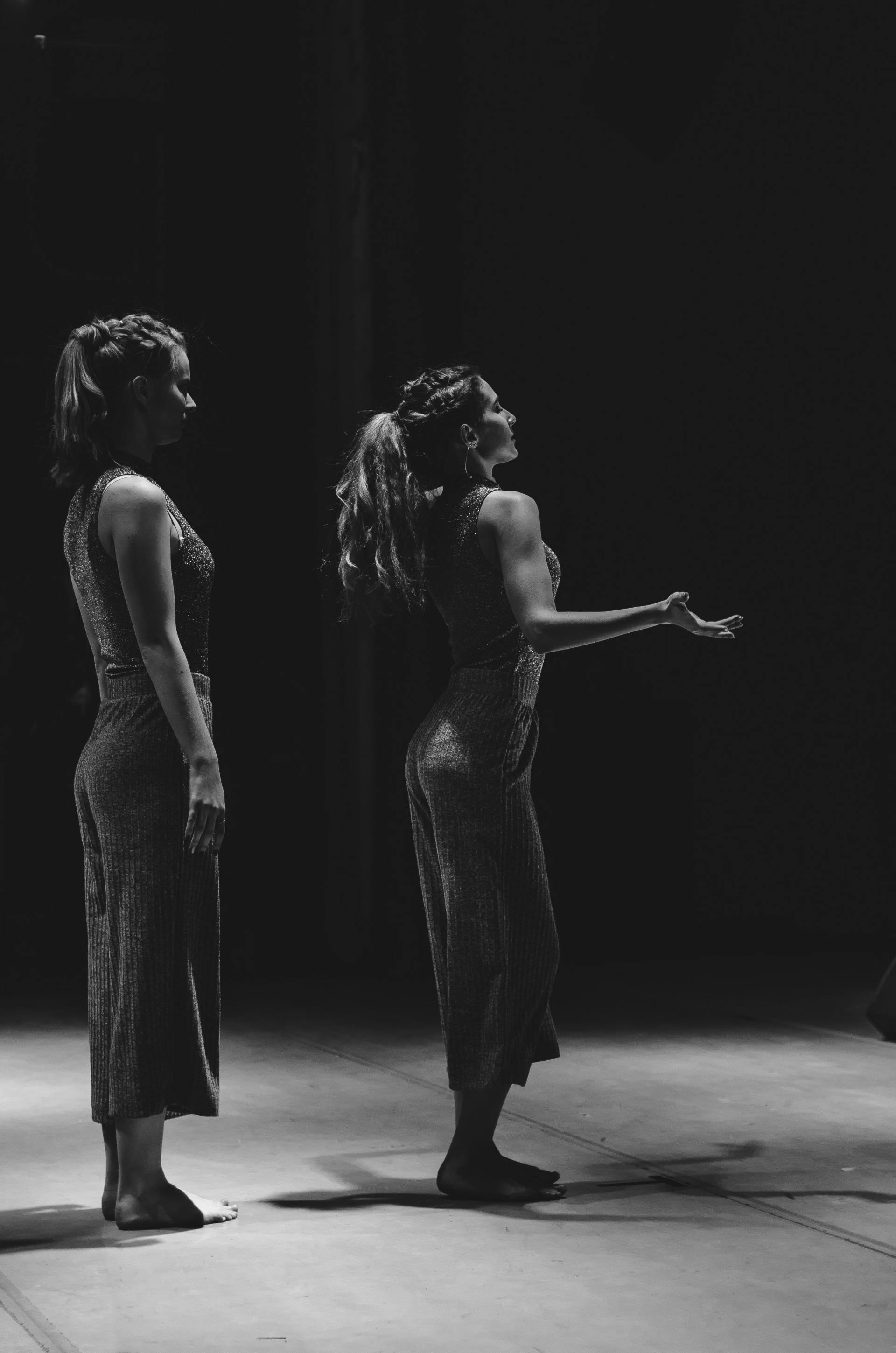 Two dancers on a stage, lit from the side. One looks upward, arms outstretched. The other stands still.