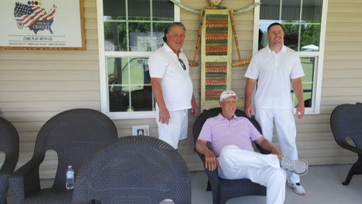Three men in white outfits pose on a porch. One seated, two standing near windows, US flag art visible.