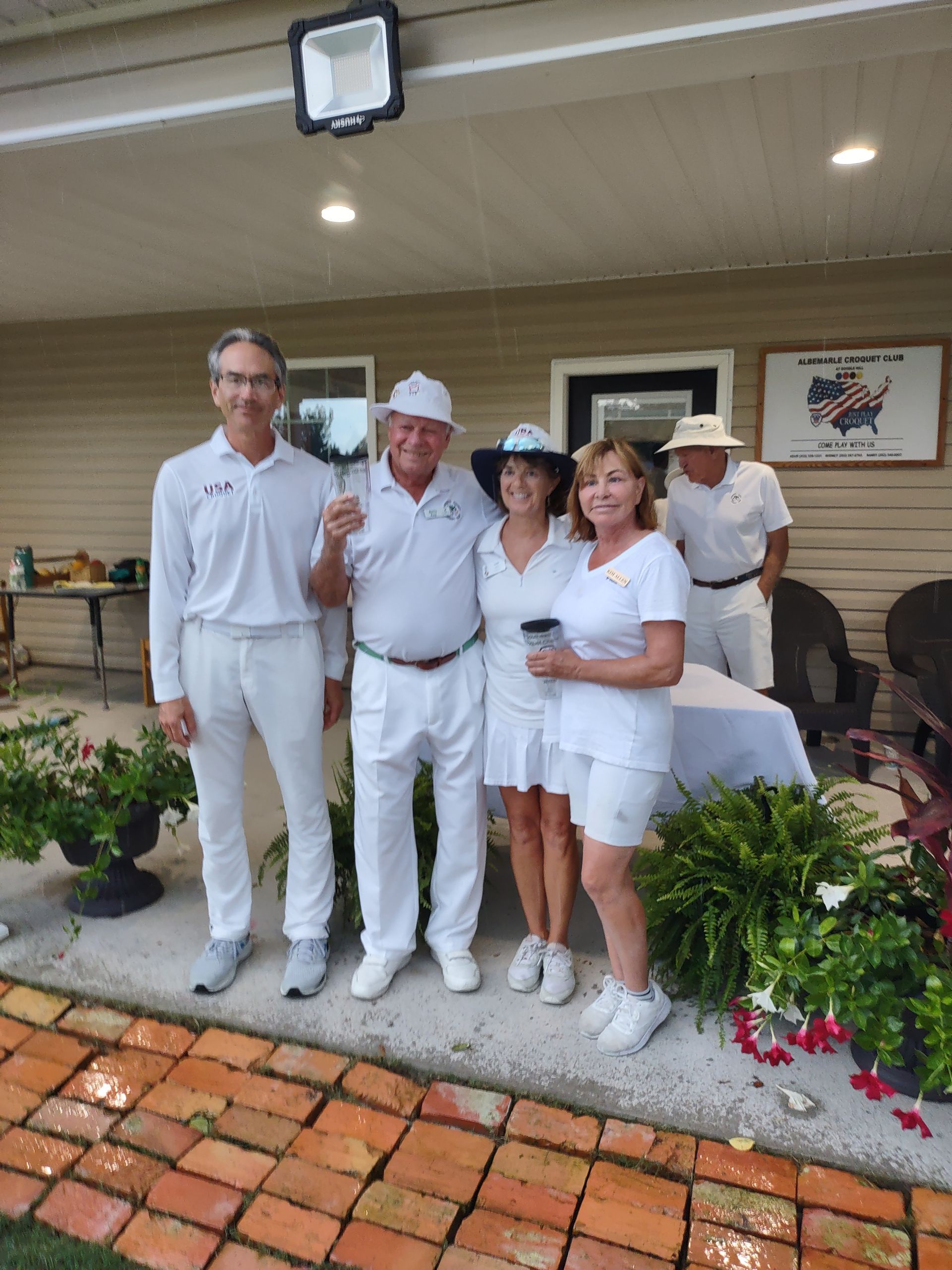 Group of five people in white attire, posing near a table, under a porch, likely at an outdoor event.