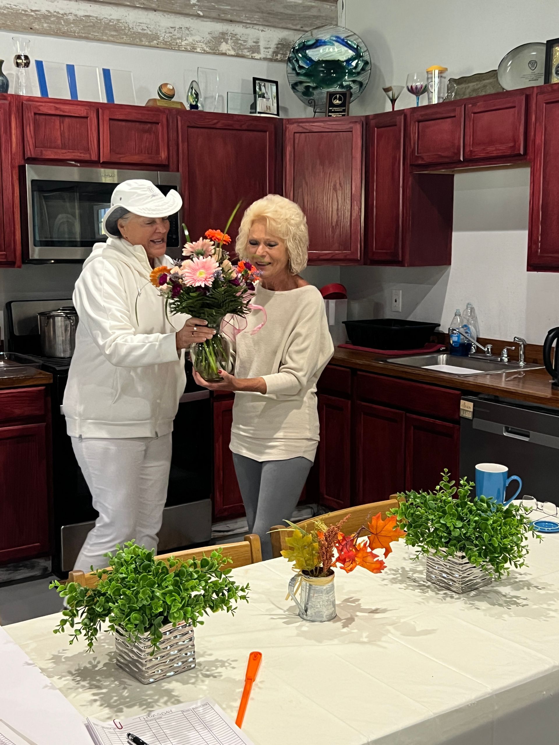Two women in kitchen; one gives flowers to the other. Burgundy cabinets, table with plants.